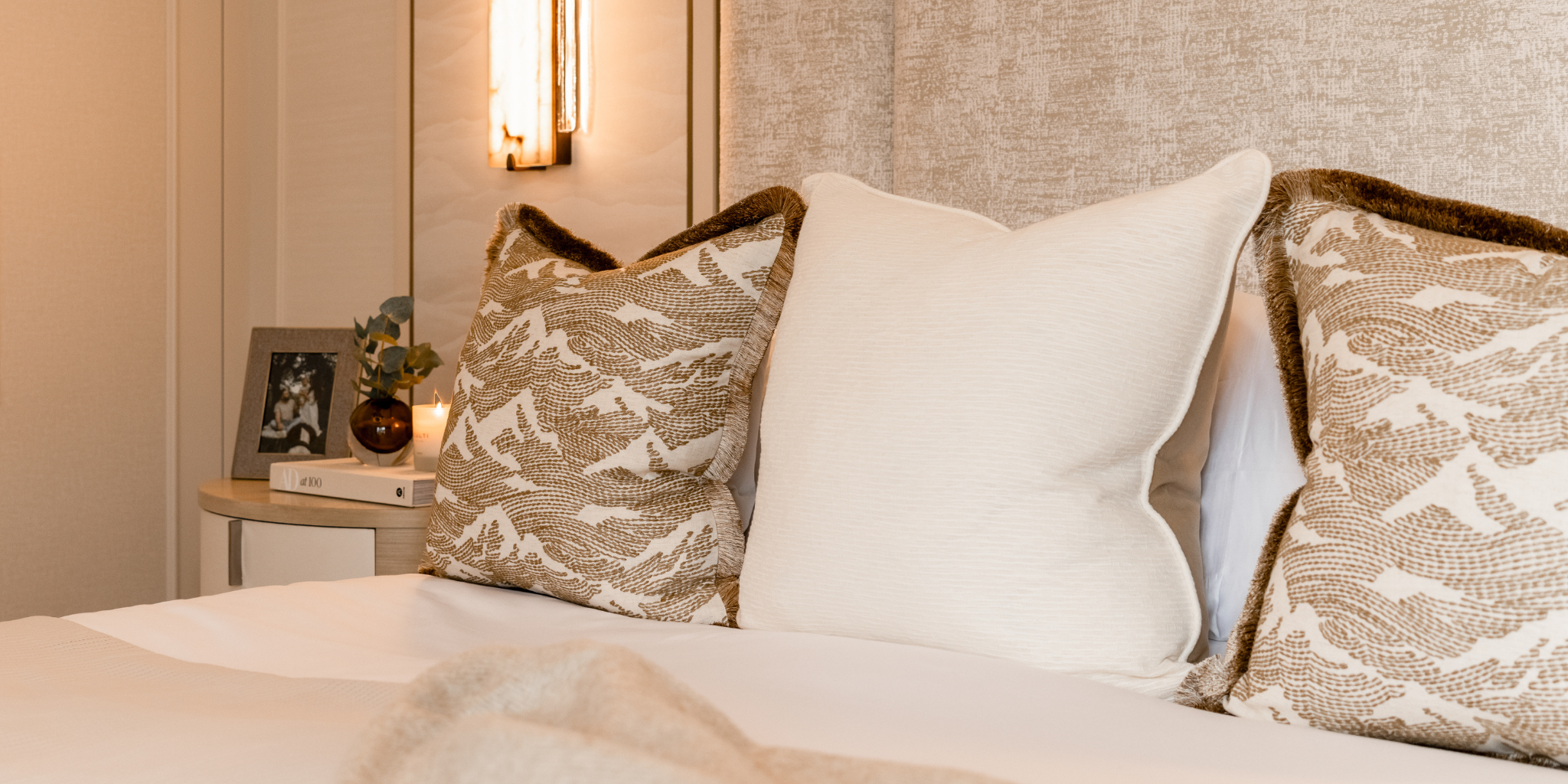 Decorative neutral pillows layered on an elegant bed with a neutral colour scheme and ambient lighting in the background.