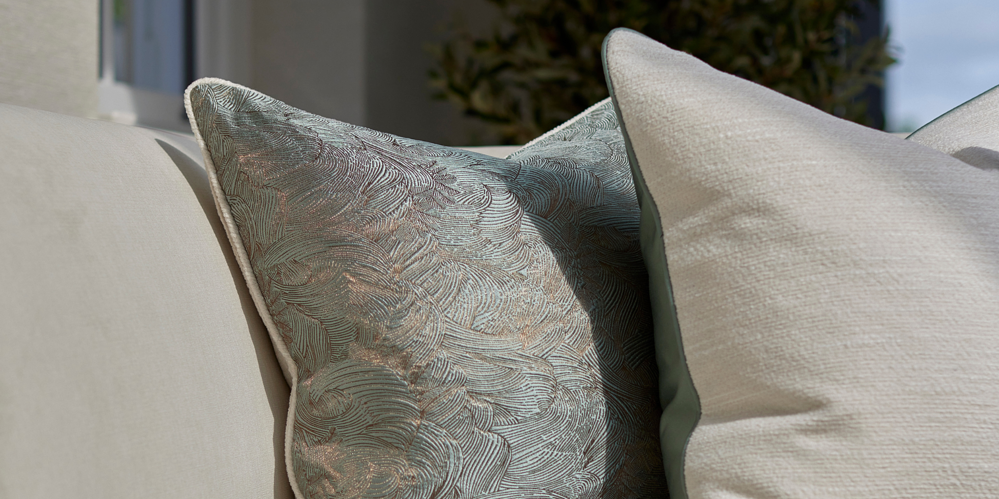 Two complementary cushions with ivory bronze and muted teal tones, layered together on a cream sofa cast with dappled natural sunlight.