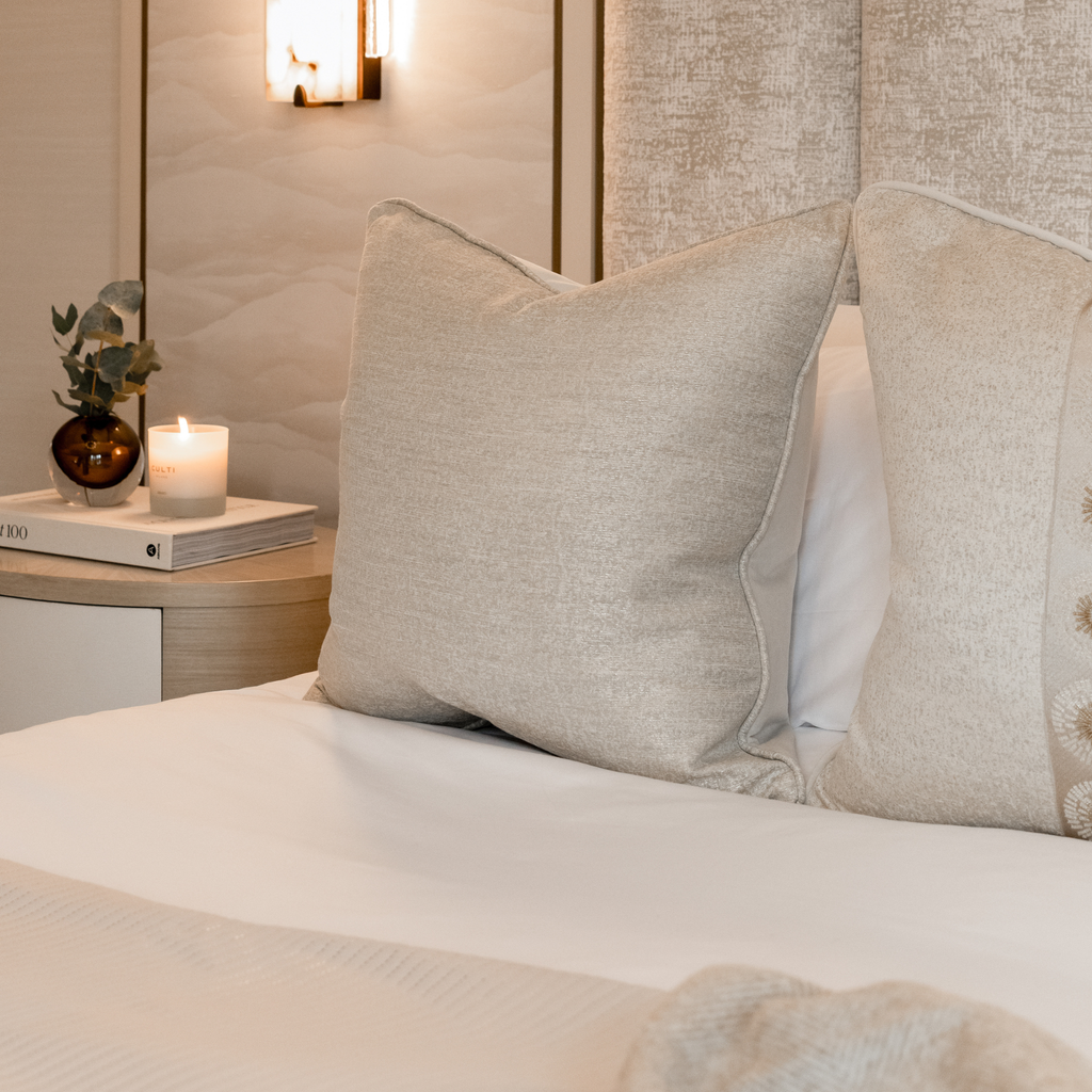 Neatly made bed with decorative neutral cushions and a candle lit in the background on a nightstand.