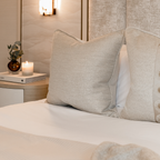 Neatly made bed with decorative neutral cushions and a candle lit in the background on a nightstand.