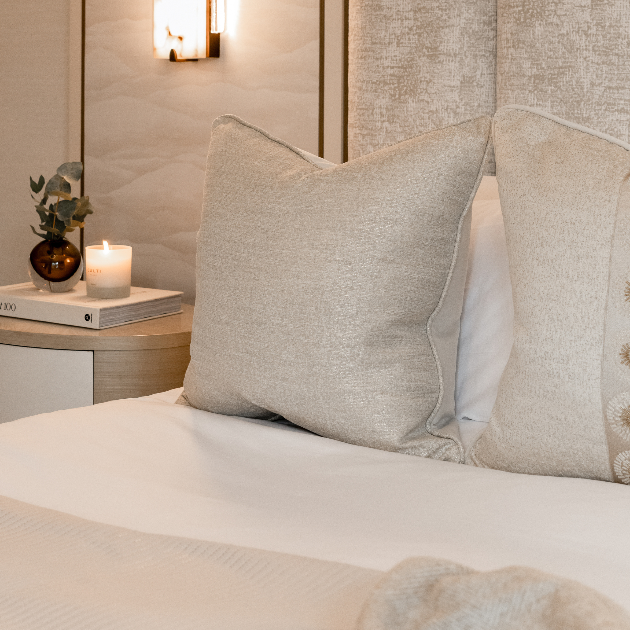 Neatly made bed with decorative neutral cushions and a candle lit in the background on a nightstand.