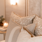 Close-up of 4 decorative neutral cushions layered together on a bed with a soft, warm background.