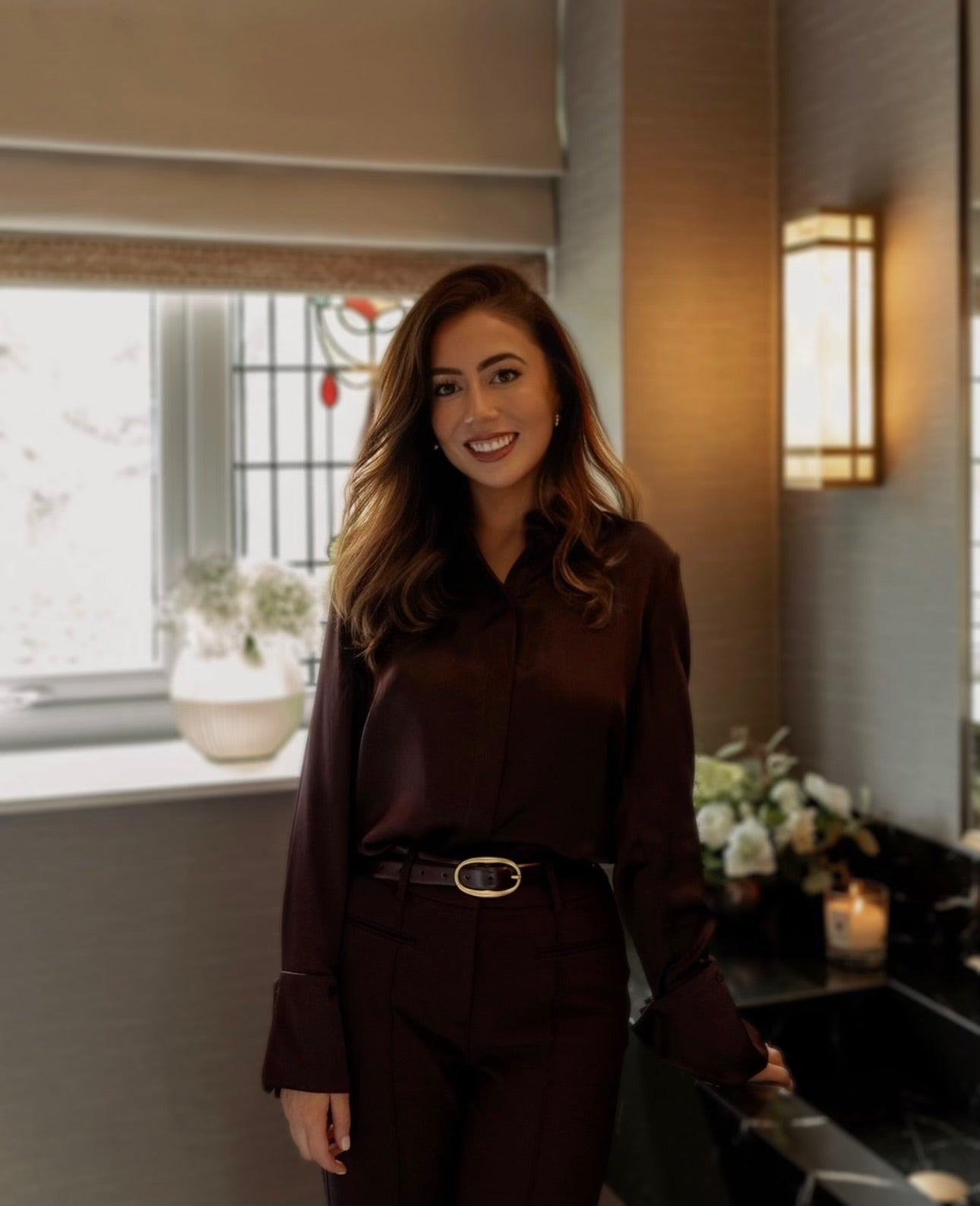 Founder Rachael Lauren stood in a tailored dark outfit, smiling at the camera whilst standing in a modern house with ambient lighting.